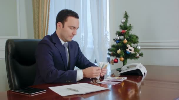 Happy young businessman counts salary near New Year tree in office — Stock Video