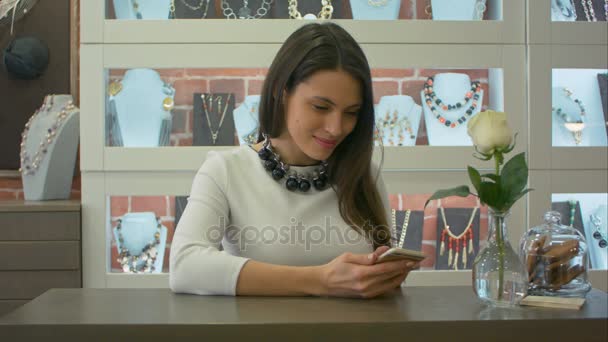 Trevlig kvinnlig säljaren skicka SMS meddelanden på smartphone och söt leende i en smyckesbutik — Stockvideo