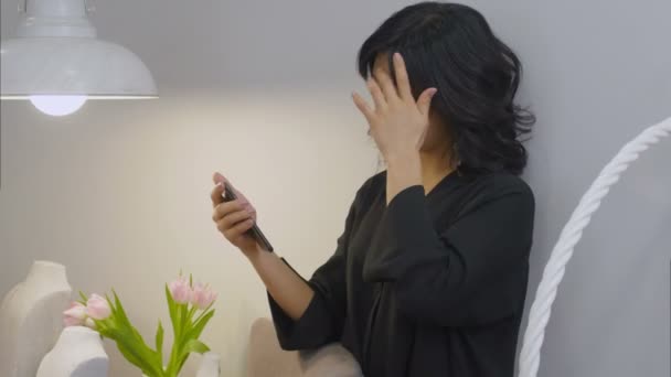 Asiatin blickt auf Smartphone-Bildschirm und lächelt im Wohnzimmer — Stockvideo