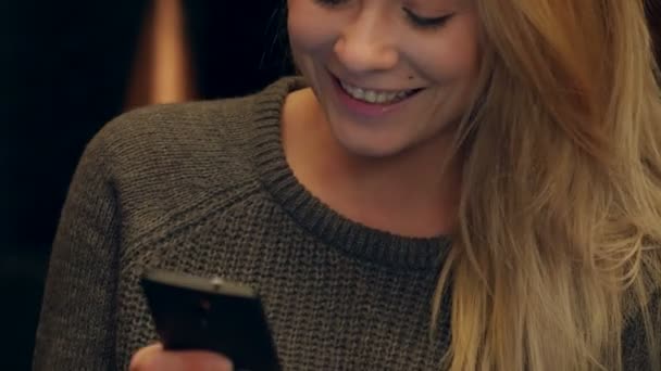 Mujer feliz usando mensajes de texto de teléfono móvil y sonriendo dentro de la cafetería por la noche — Vídeo de stock