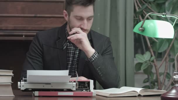 Joven serio con gafas sentado en el escritorio con máquina de escribir y libro de lectura — Vídeo de stock