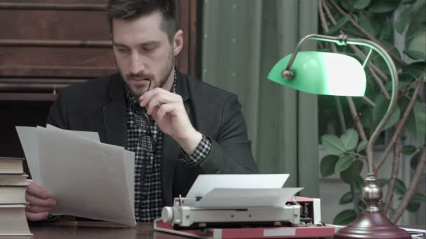 Young journalist reading printed articles sitting at the desk with typewriter — Stock Video