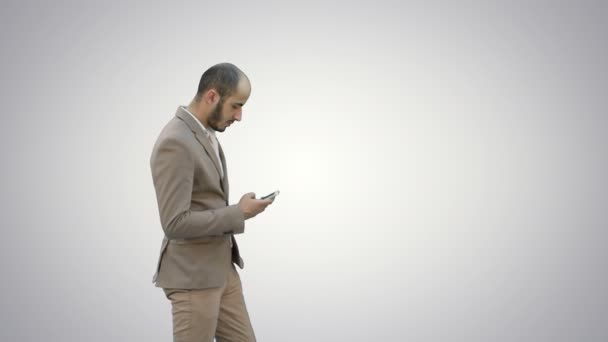 Hombre joven en traje caminando y enviando mensajes de texto en el teléfono móvil sobre fondo blanco . — Vídeo de stock