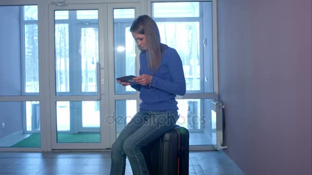 Жінка використовує цифровий планшет, сидячи на валізі у вестибюлі — стокове відео