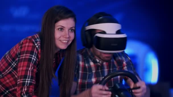 Bella ragazza sorridente guardando il suo ragazzo che gioca al videogioco di corse in cuffia realtà virtuale — Video Stock