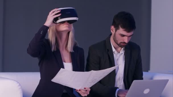 Donna bionda in VR auricolare tenendo piani di costruzione mentre il suo partner maschile digitando sul computer portatile — Video Stock