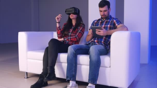 Pareja de alta tecnología: chica que usa un dispositivo de realidad virtual mientras un joven escribe algo en su tableta digital — Vídeo de stock