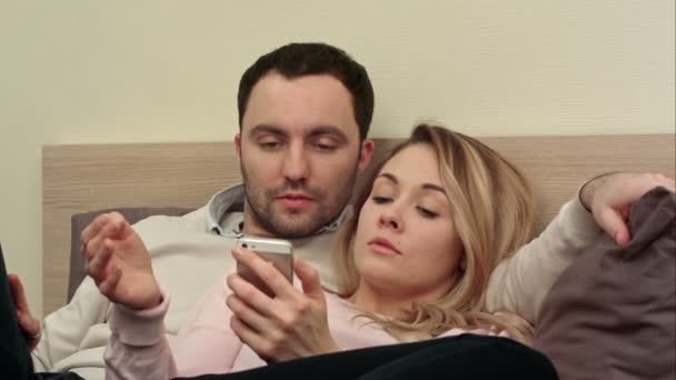 美しいカップルはベッドに座って、スマート フォンは、画面に触れる旅行から写真を探して — ストック動画