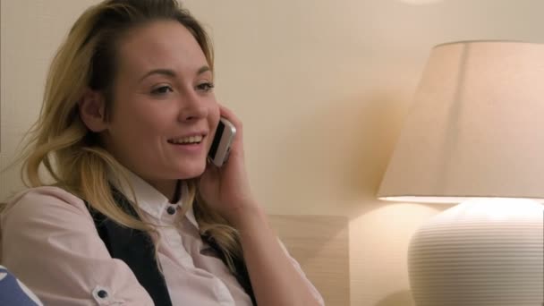 Молодая женщина имеет положительный разговор с помощью мобильного телефона сидя на кровати — стоковое видео