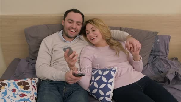 Hermosa pareja sentada en la cama y mirando la foto del viaje en el teléfono inteligente, tocando la pantalla, sonriendo — Vídeo de stock