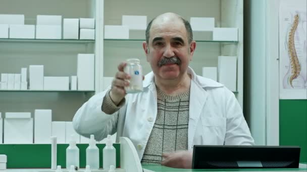 Médico sonriente sostiene un frasco de tabletas o pastillas con una etiqueta blanca en blanco para el tratamiento de una enfermedad y mira a una cámara — Vídeos de Stock