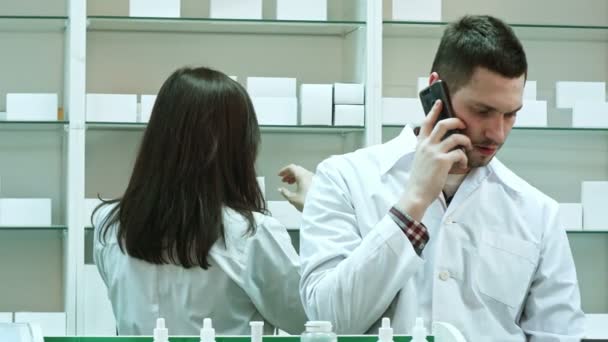 Vrouwelijke en mannelijke apothekers werken in de apotheek, praten via slimme telefoon en controleren van pillen — Stockvideo
