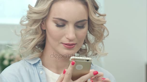 Blondes Mädchen sitzt im Wohnzimmer und blickt lächelnd auf Smartphone-Bildschirm — Stockvideo