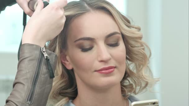 Junge hübsche Frau benutzt Smartphone im Schönheitssalon, während Friseur die Frisur macht — Stockvideo