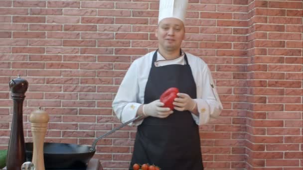 Chef de mediana edad en uniforme blanco de pie cerca del escritorio, hablando con la cámara con vegetales en las manos, listo para preparar una ensalada — Vídeo de stock