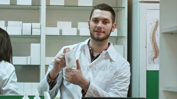 Joven malefarmacista sosteniendo un frasco blanco de pastillas, promoviendo la medicina, mientras su colega trabaja — Vídeo de stock