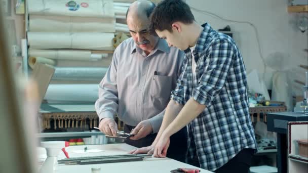 Giovane tirocinante che studia come costruire una cornice, lavoratore anziano parlando con lui dietro la scrivania in laboratorio cornice — Video Stock