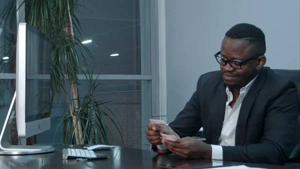 Afro businessman counting cash, sitting at desk in office