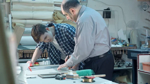 Trabajador de edad avanzada instruyendo a los jóvenes aprendices cómo cortar un vidrio para el marco detrás del escritorio en el taller de marco — Foto de Stock