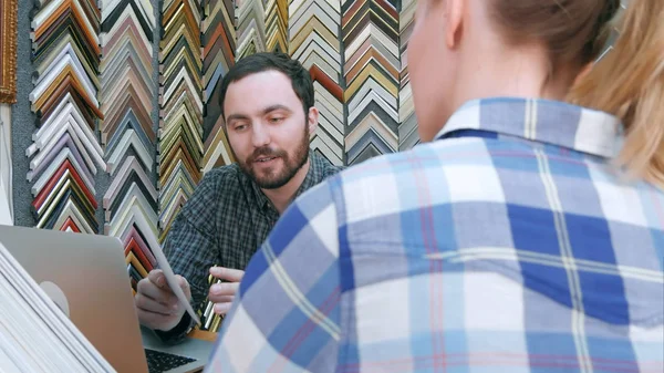 Trabajador joven charlando con el cliente sobre los detalles del marco de la imagen, utilizando el ordenador portátil en el mostrador en el taller — Foto de Stock