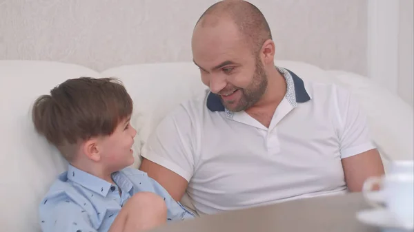 Sonriente padre hablando con su hijo sentado en el sofá blanco y bebiendo té —  Fotos de Stock