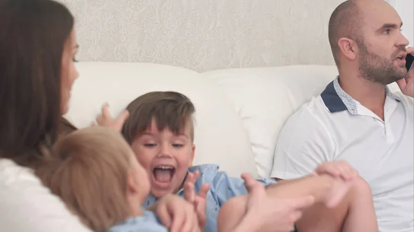 Отец разговаривает по телефону, пока маленькие мальчики играют со своей матерью на диване — стоковое фото