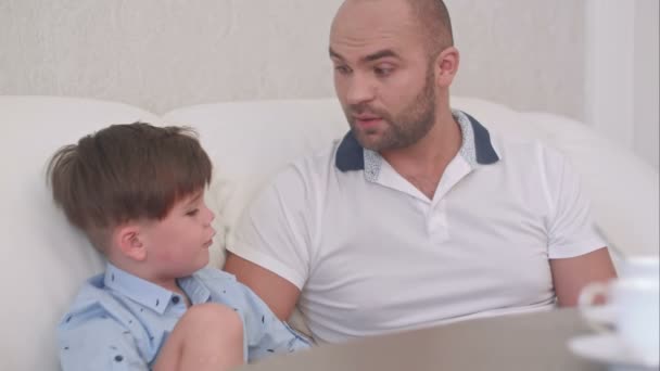 Sonriente padre hablando con su hijo sentado en el sofá blanco y bebiendo té — Vídeo de stock