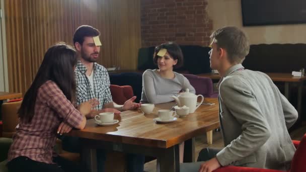 Молодые красивые люди за столом кафе играют в именную игру со стикерами, приклеенными к их лбу — стоковое видео