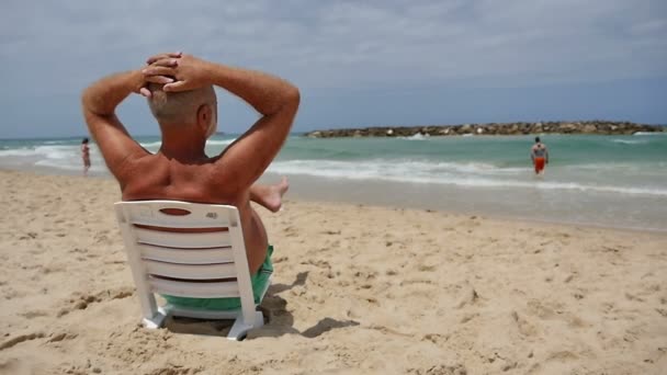 Зрелый мужчина сидит на пляжном стуле и смотрит на море, наслаждаясь отдыхом — стоковое видео