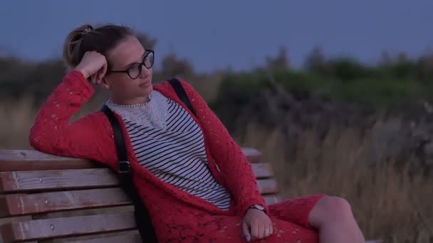 Mädchen mit Brille sitzt auf Bank und beobachtet das Abendmeer — Stockvideo