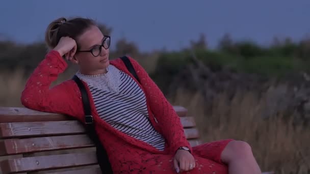 Fille dans des lunettes assis sur le banc regarder la mer du soir — Video