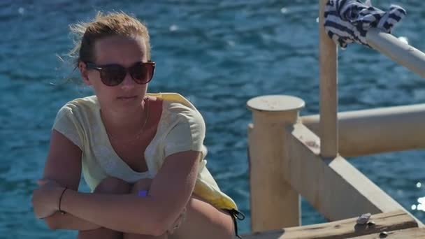 Young girl in sunglasses sitting near the seashore on a sunny day — Stock Video
