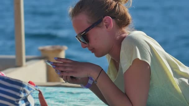 Mujer joven en gafas de sol acostada con smartphone en una playa — Vídeos de Stock