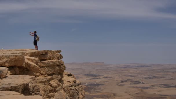 Молодой человек стоит на краю скалы и делает панорамное фото пустыни на телефон — стоковое видео