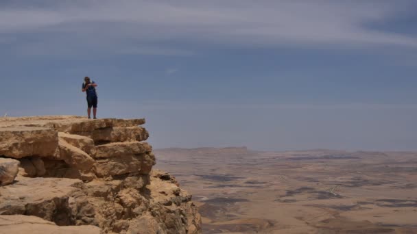 Молодой человек стоит на краю скалы и фотографирует пустыню на телефон. — стоковое видео
