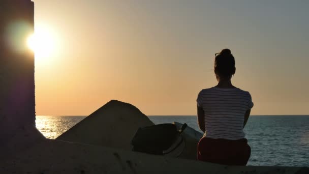 Молодая женщина сидит на скалах и смотрит на закат моря — стоковое видео