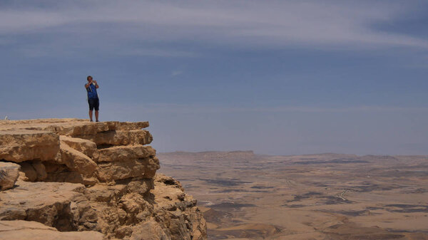 Young man standing on cliff edge and taking pictures of the desert on his phone