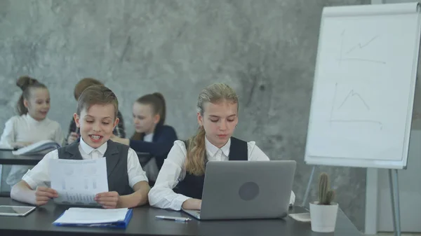 Двое маленьких детей в деловых костюмах сидят за столом с ноутбуком и выглядят счастливыми — стоковое фото