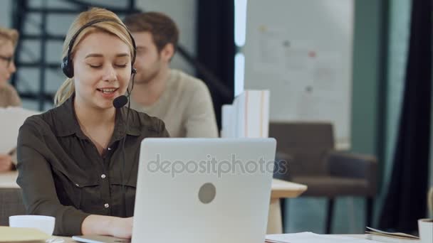 Sorrindo operadora de linha de ajuda feminina com fones de ouvido em sua mesa no escritório — Vídeo de Stock