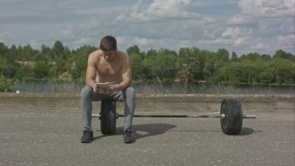 Молодий білий спортсмен використовує свій планшетний комп'ютер після поїзду на відкритому повітрі — стокове відео