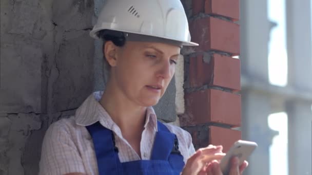 Una joven trabajadora de la construcción se sienta en un sitio y trabaja en un teléfono inteligente — Vídeos de Stock