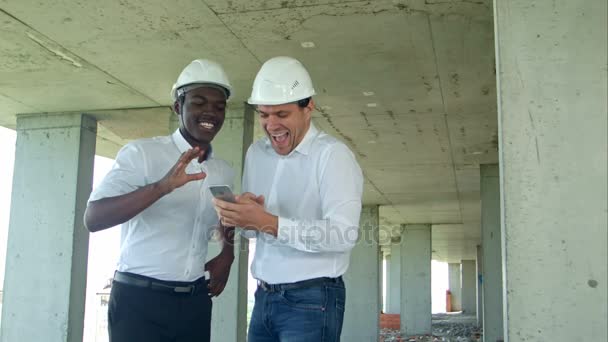 Constructores de construcción sonriendo mientras se utiliza el teléfono inteligente en el sitio — Vídeos de Stock