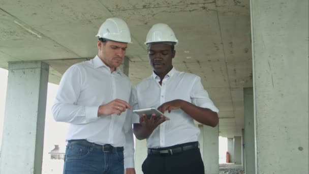 Африканский американский инженер и кавказский архитектор с использованием цифровых планшетов и в защитных шлемах на строительной площадке — стоковое видео