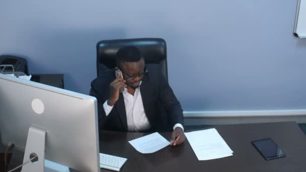 Jeune homme d'affaires afro-américain pensif ayant un appel téléphonique, discutant de documents et siégeant au bureau — Video