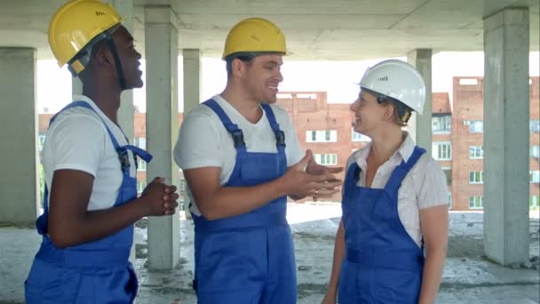 Группа улыбающихся строителей в хардхатах разговаривает на стройке — стоковое видео