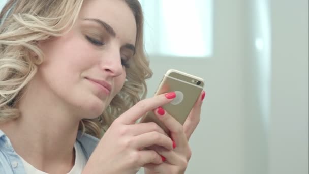Feliz joven rubia está sentada en un salón de belleza y usando un teléfono inteligente — Vídeos de Stock
