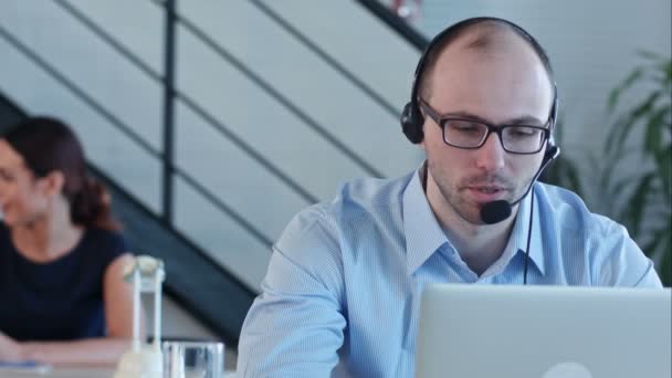 Joyful call center agent met zijn headset praten op zoek naar laptop — Stockvideo