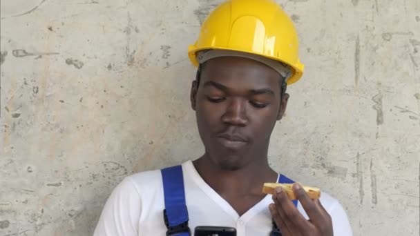 Trabalhador afro-americano faminto no canteiro de obras comendo comida sanduíche almoço olhando para smartphone — Vídeo de Stock