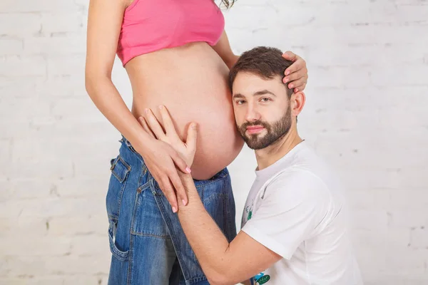 Bonito homem está ouvindo a barriga de sua bela esposa grávida — Fotografia de Stock