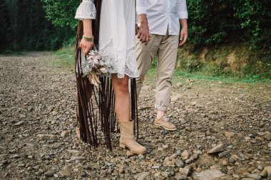 Gelin ve damat bir dağ yolundalar. Bir adam ve bir kadın kayalık bir yolda rahat ayakkabılarla yürüyorlar. Gelin çiçekleri elinde tutuyor..